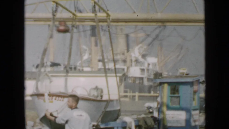 The small boat remains suspended near a dock worker, with fishing boat rigging, a blue booth, and harbor equipment behind it.
