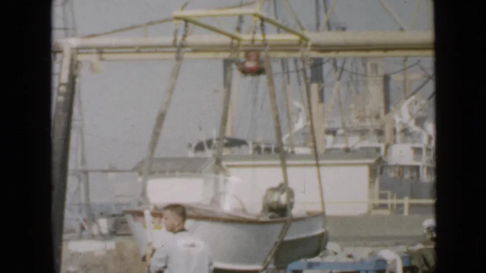 A small boat hangs in slings below the gantry crane while a man stands on the dock, with a larger fishing vessel behind.