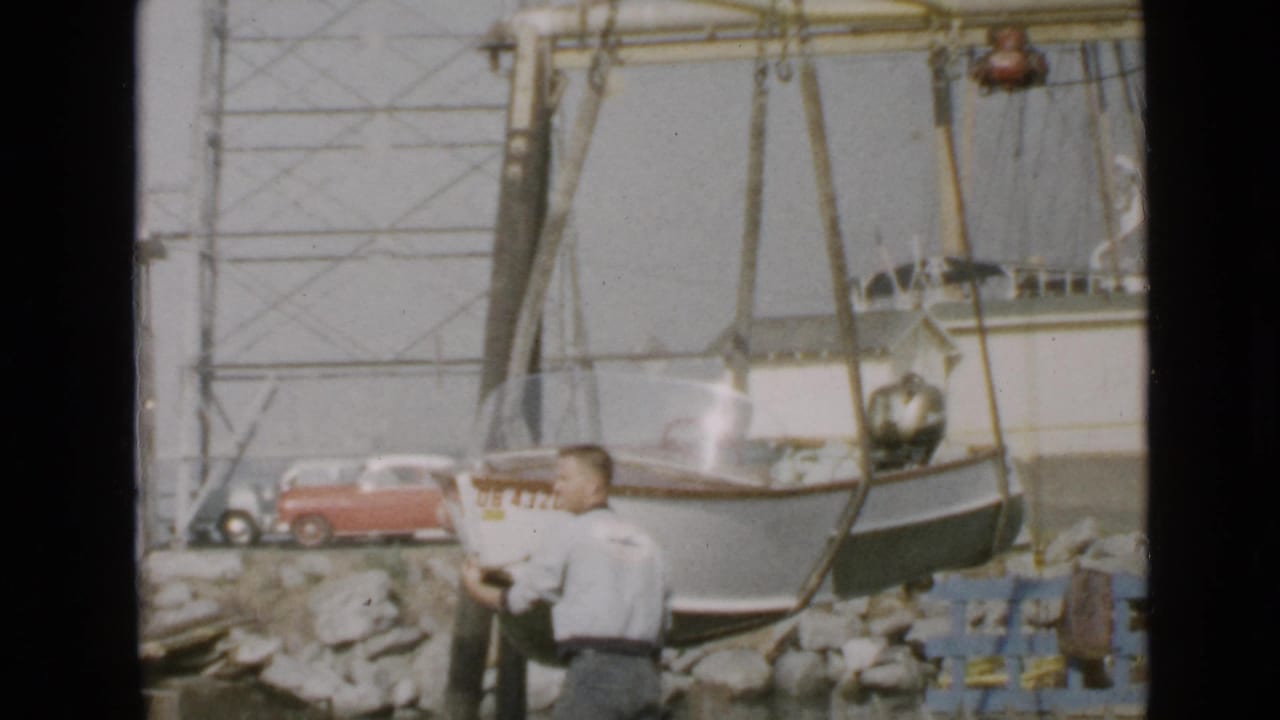 Man standing near a small white boat being lifted beside rocks and a red car.