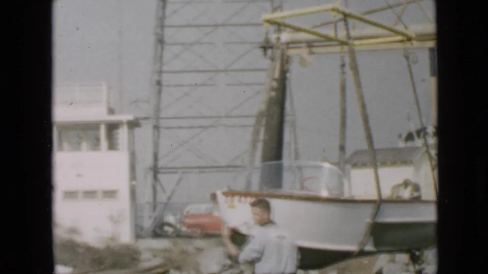 A small numbered boat is suspended by straps near a white building and red car in San Diego waterfront footage from 1949.