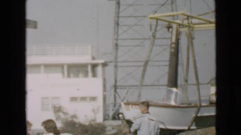 Faded 8mm frame of a small boat under crane rigging with a white building, tower, and people partly visible at the lower edge.