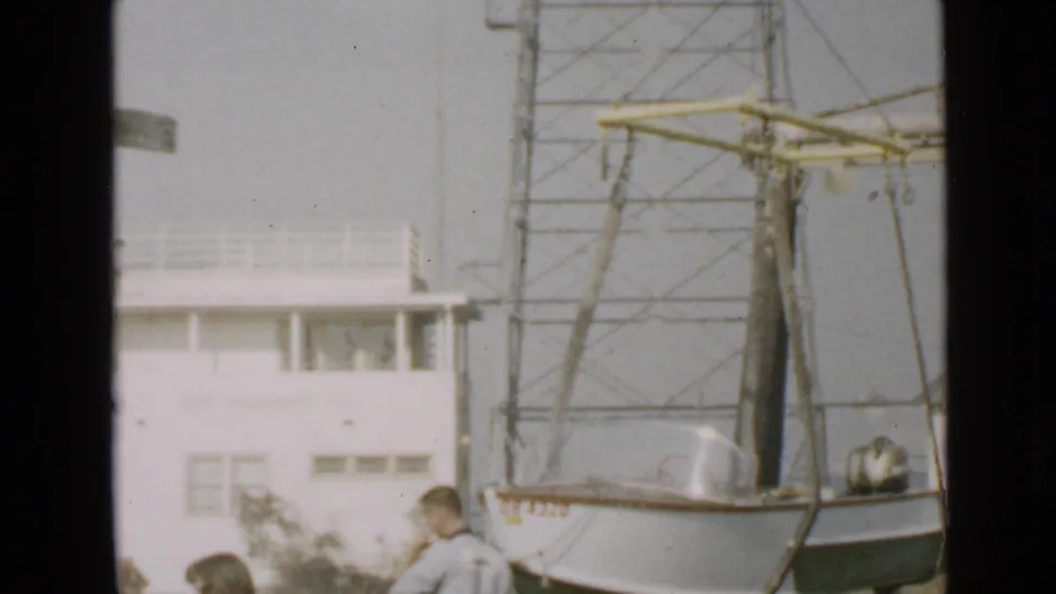 San Diego 1949 frame showing a man near the ground as a small white boat remains suspended beside crane rigging and a white building.