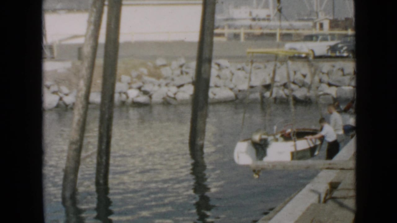 Two men position a small boat beside a wooden dock with pilings and water in view.