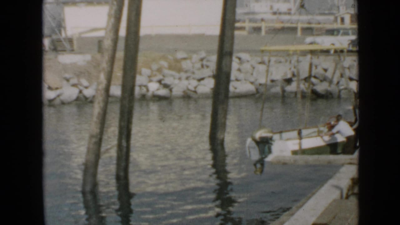 A small boat sits beside the dock as two men work near tall wooden pilings.