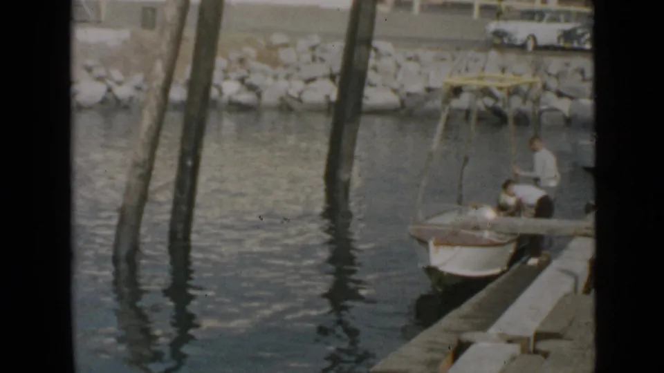 A 1949 San Diego home-movie frame shows the men closer to the dock edge as the boat rests in the water near pilings and rocks.