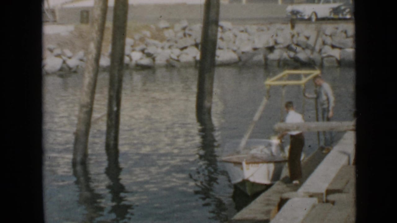 Two men stand on a wooden dock beside a small motorboat in calm harbor water.