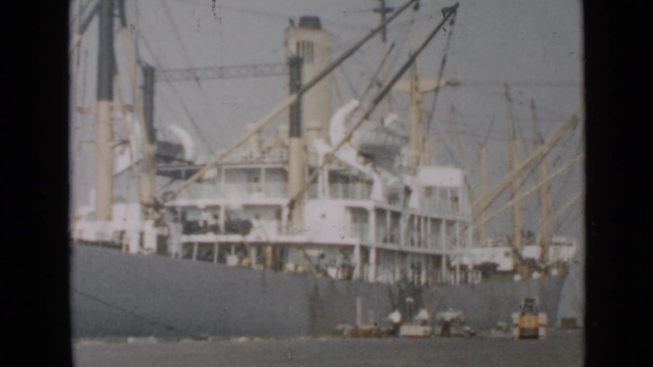 Large cargo ship docked at a harbor with masts, rigging, and small figures near the hull.