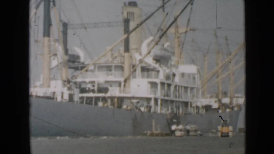 1949 San Diego harbor home-movie frame centered on a large cargo ship, its masts, rigging, hull, and small dockside activity.