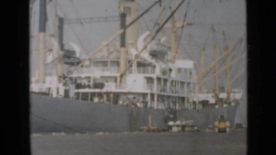 1949 San Diego harbor frame showing a large docked cargo ship with rigging, visible hull, small people near the dock, and a small vessel nearby.