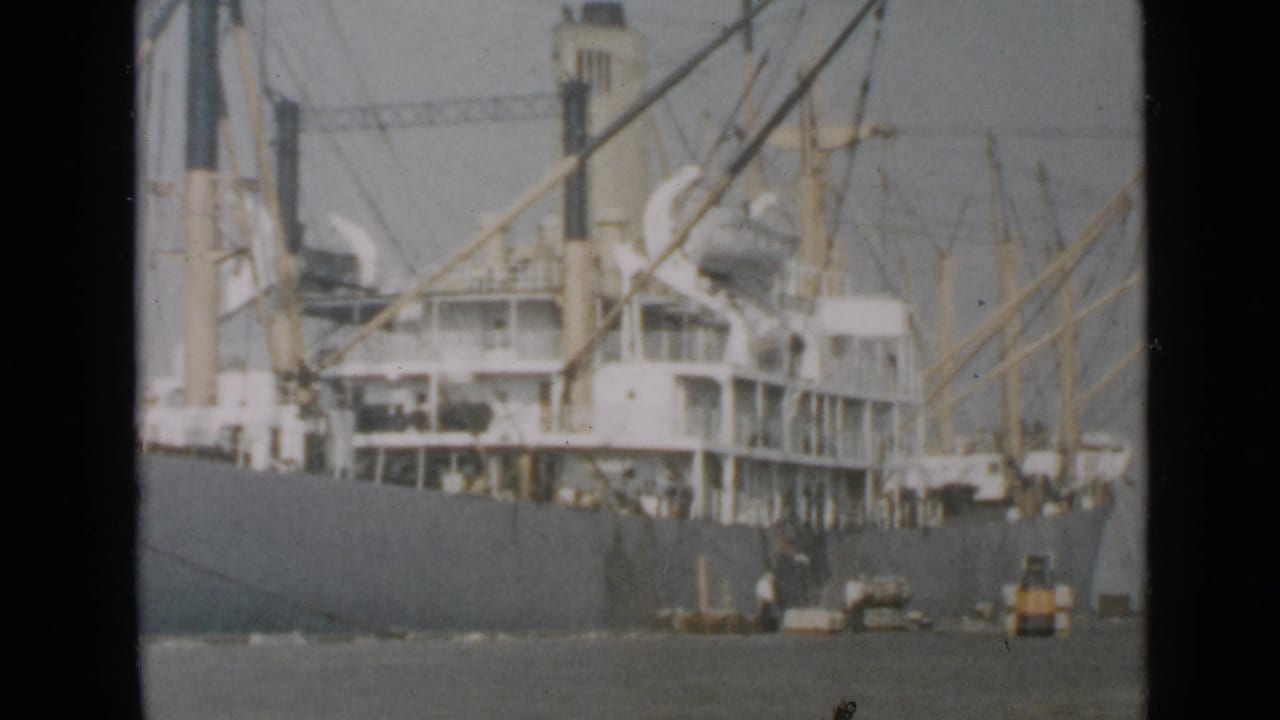 The docked 1949 San Diego cargo ship remains in view, with rigging, cranes, workers, and a yellow forklift-like vehicle near the hull.