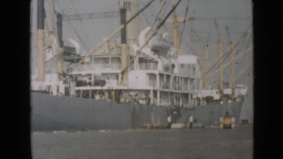 Soft 8mm frame of the San Diego cargo ship's side, showing cranes above the deck and workers clustered near equipment on the pier.