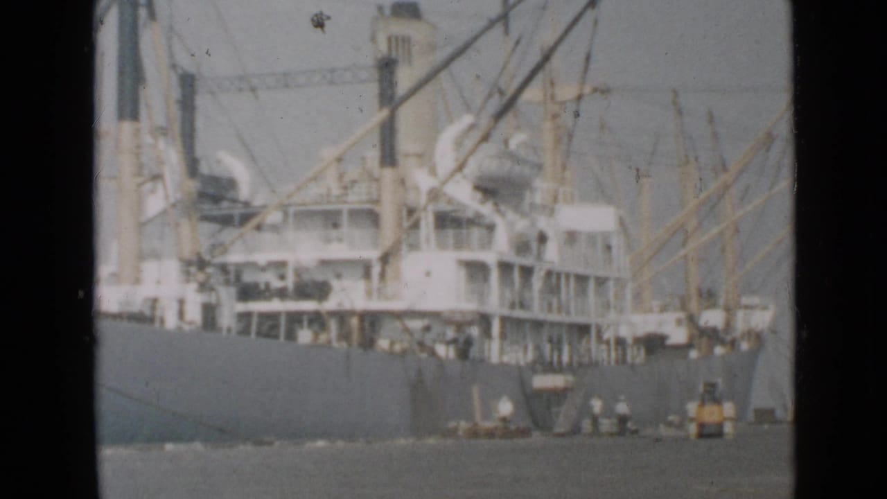 Large cargo ship at a harbor with cranes, rigging, small boats, and hazy water.