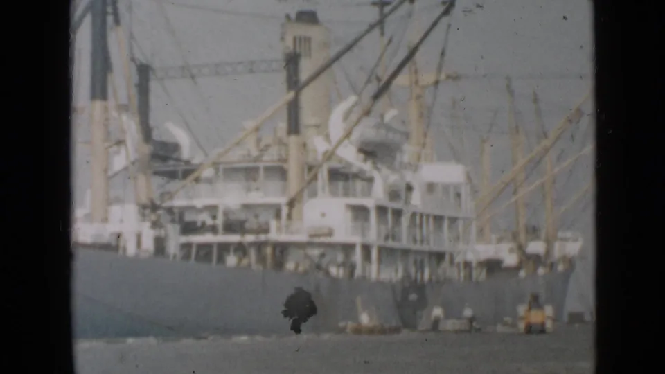 Large cargo vessel in San Diego harbor with visible cranes or booms, rigging, small craft, and aged 8mm film marks.