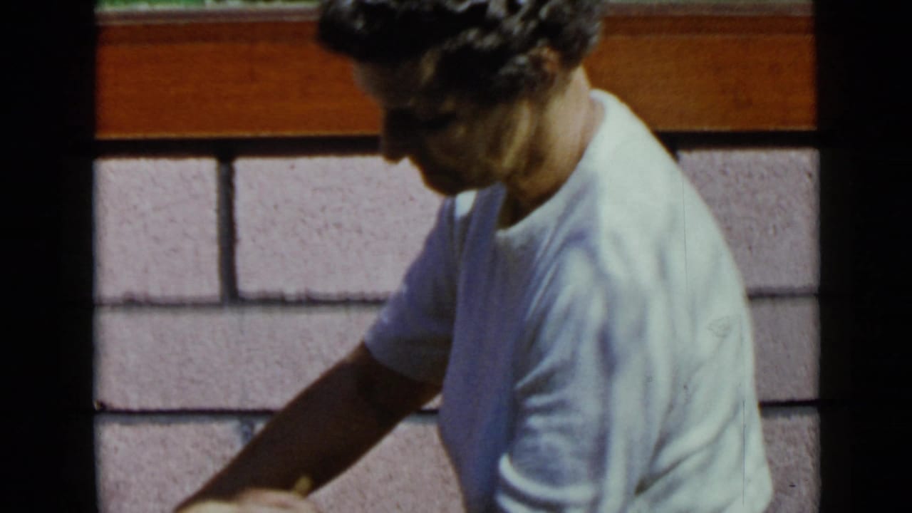 In 1949 San Diego home-movie footage, a curly-haired man in a light T-shirt bends near a pink cinder block wall.