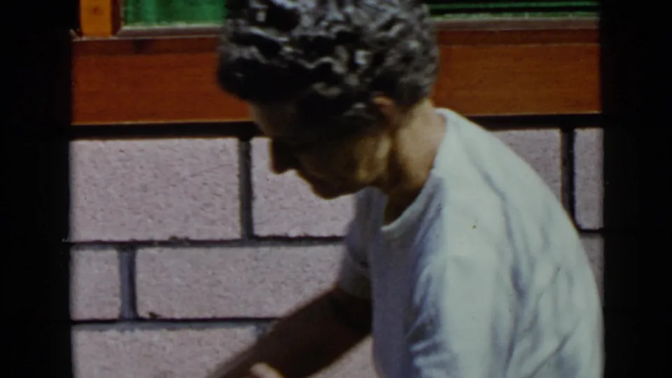 A man in a light T-shirt bends forward with his arm extended beside the pink wall in San Diego home-movie footage.