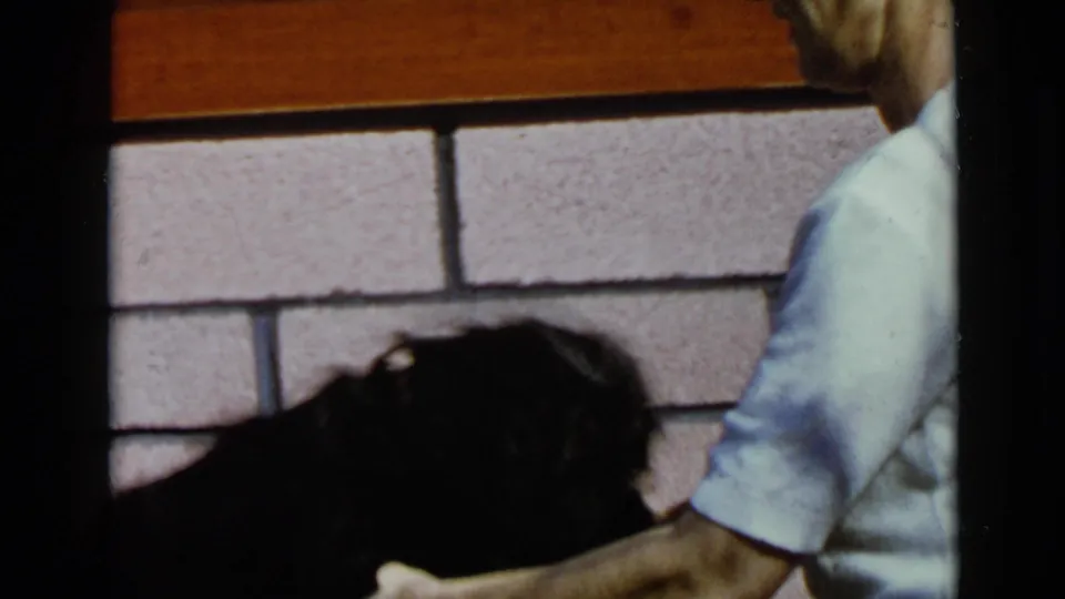 1949 San Diego home-movie frame showing a black poodle beside a partial adult figure near a block wall.