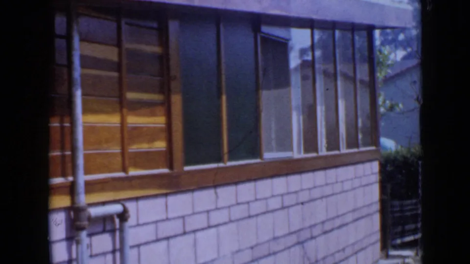 Mid-century San Diego residential exterior with a row of wood-framed windows above pale shingle siding in 8mm footage.