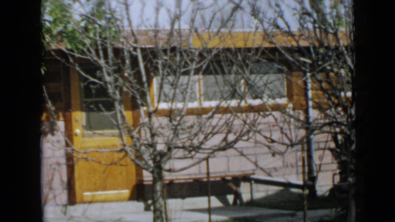 1949 San Diego home-movie frame showing a house exterior with an orange door, white facade, dark windows, and bare branches in front.