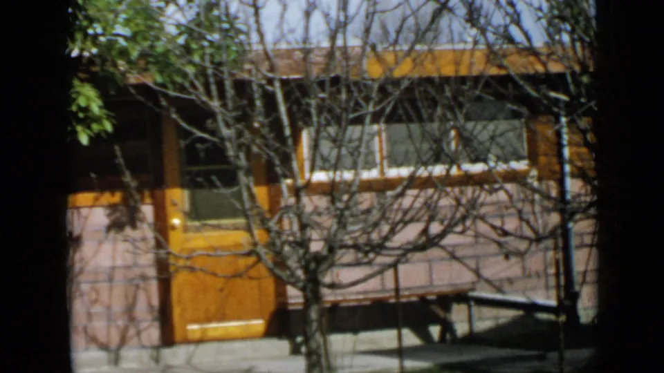 1949 San Diego frame showing the same house exterior with orange door, white facade, foreground branches, and slight handheld reframing.