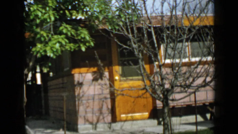1949 San Diego home-movie frame of the small house exterior, with yellow door, brick wall, windows, and branches crossing the view.