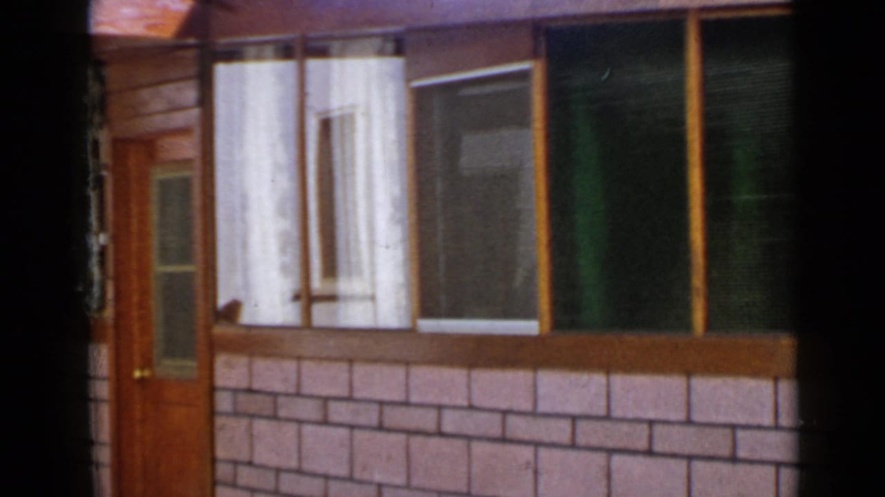 1949 San Diego home-movie frame showing a wood door, broad curtained windows, brick-pattern facade, and shaded edges.