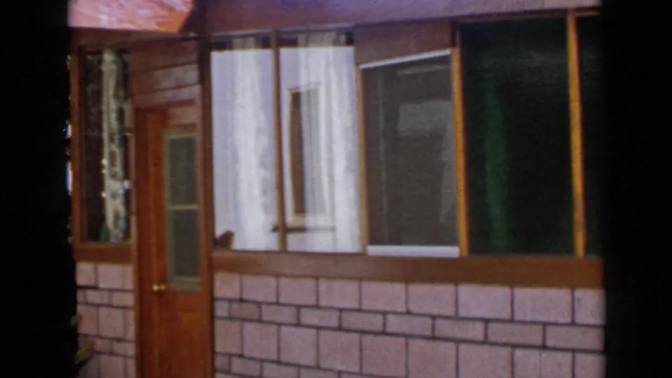 1949 San Diego home-movie still of a modest house front, with curtained windows, wood trim, a door, and dark foreground obstruction.