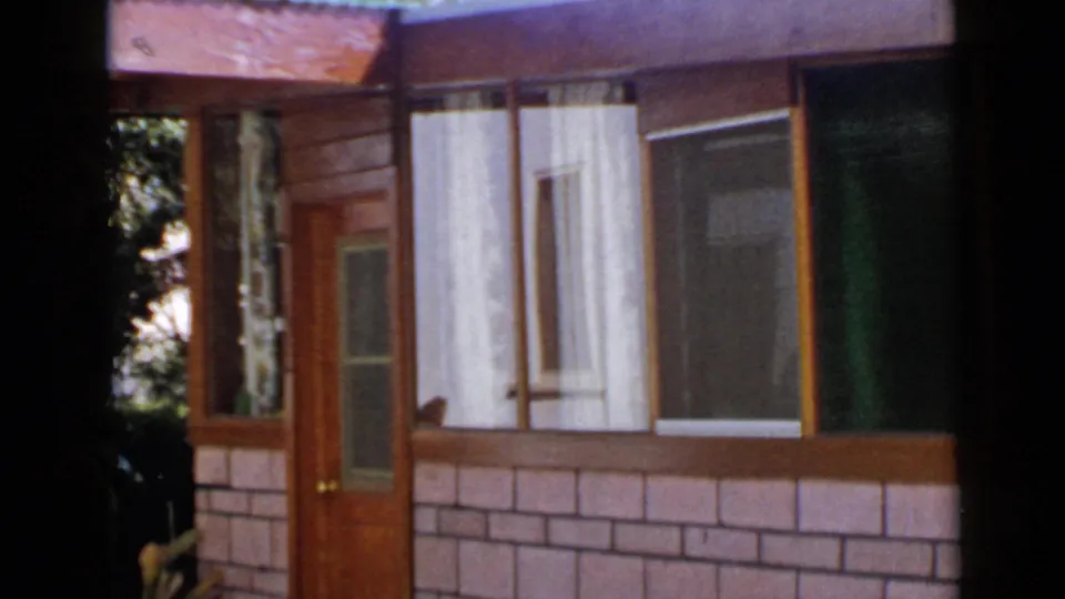 1949 San Diego frame showing the house entrance, broad curtained windows, brick-pattern exterior, and nearby dark foliage.