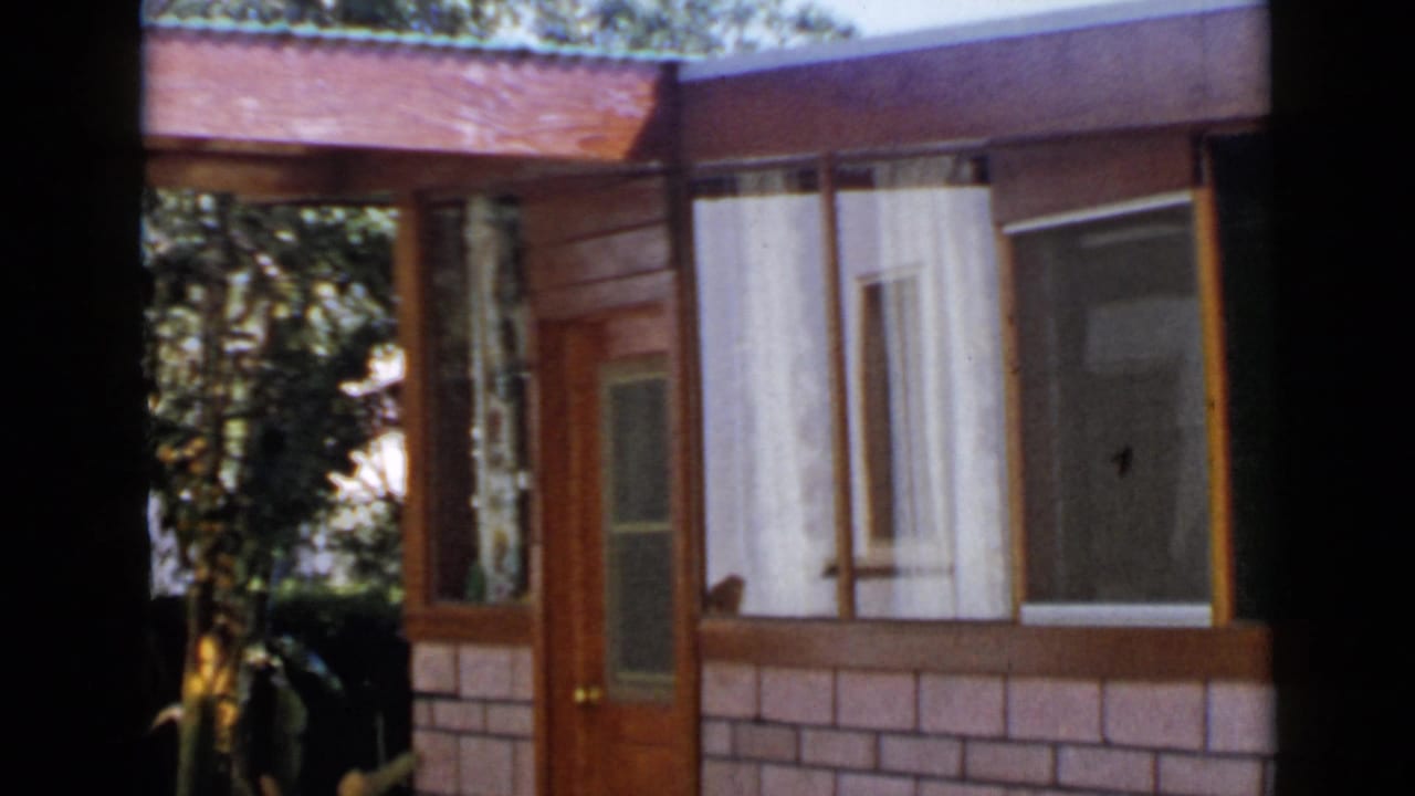 1949 San Diego 8mm frame of a house exterior with a wooden entry door, brick lower wall, sheer window curtains, and leafy plants nearby.