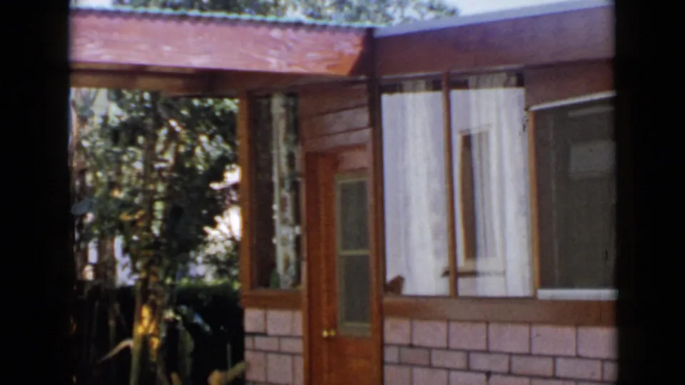 1949 San Diego home-movie frame with the house facade shifted slightly, showing the door, brick base, wood cladding, curtained windows, and shaded trees.