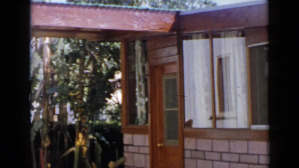 1949 San Diego 8mm frame showing the same house front with a glass-paneled door, wood siding, brick lower wall, window curtains, and garden plants.