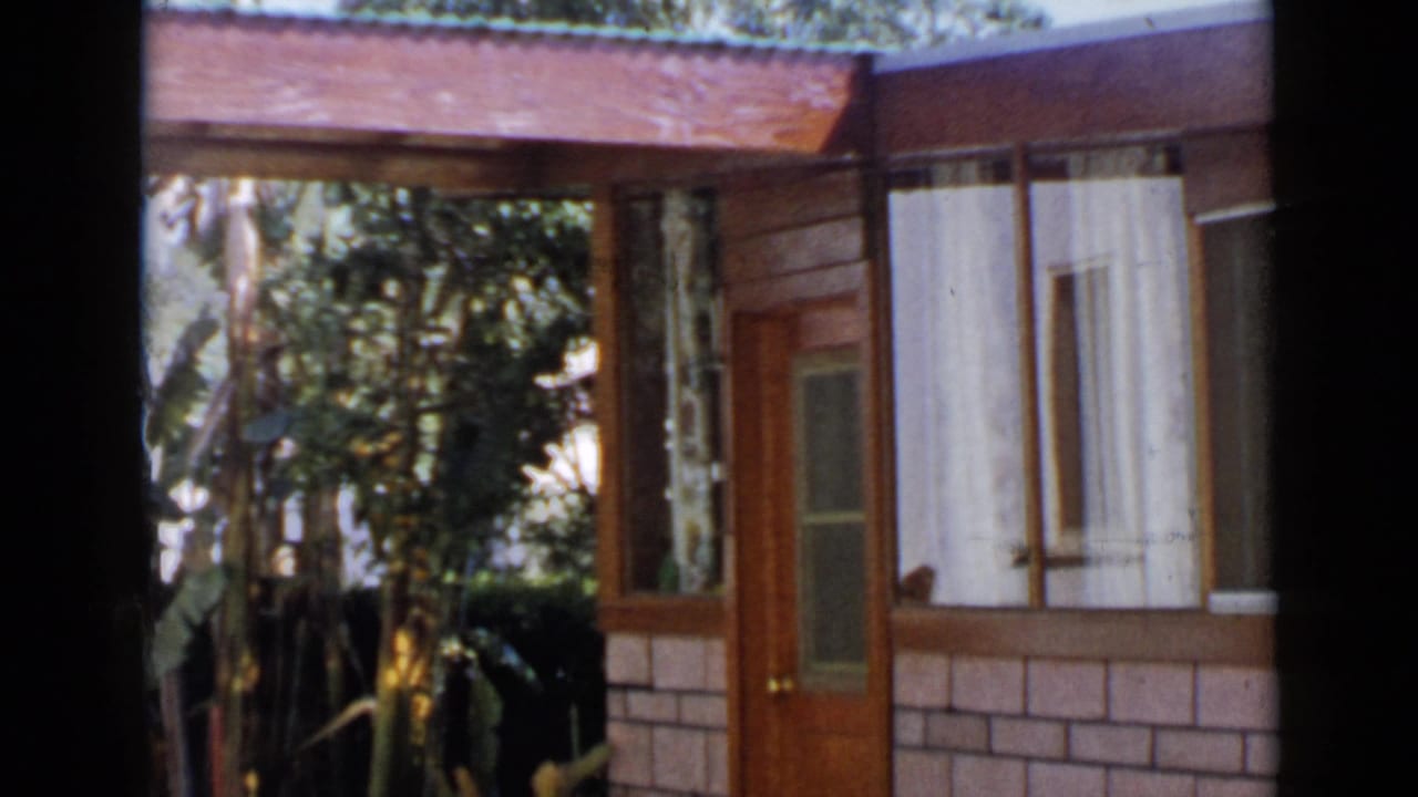 1949 San Diego home-movie frame showing a wood-and-brick house exterior with glass door, windows, and surrounding greenery.