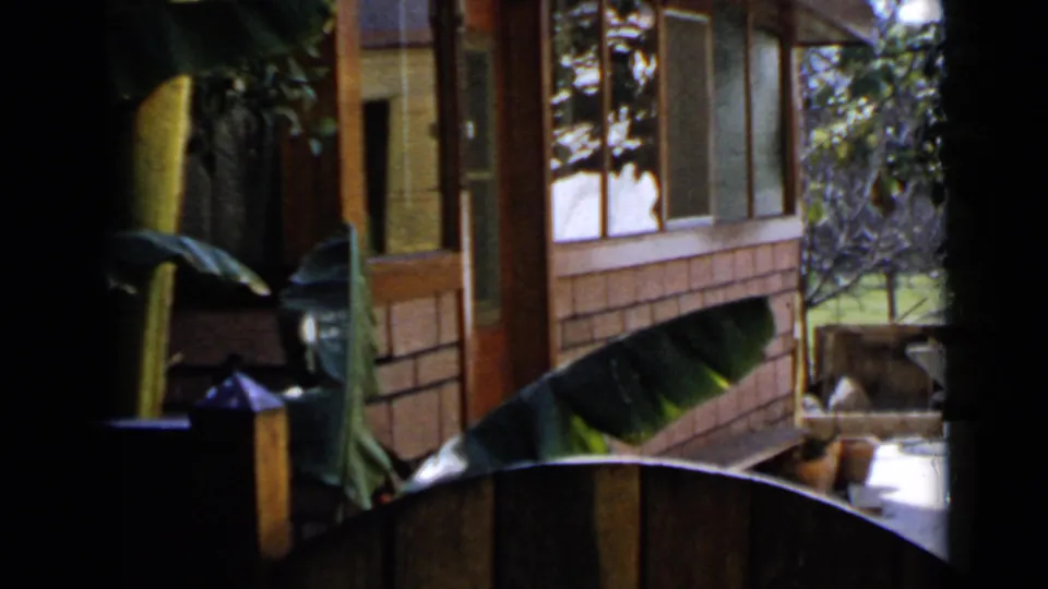 1949 San Diego frame showing the side of a wood house with porch posts, screened windows, and banana-like leaves in the foreground.