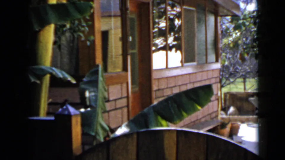 1949 San Diego home-movie view of a house side with brick accents, wood posts, screened windows, and tropical planting.