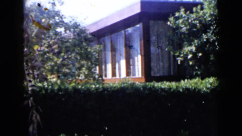1949 San Diego home-movie frame of a low modern building with tall windows, dense hedge, and leafy trees around it.