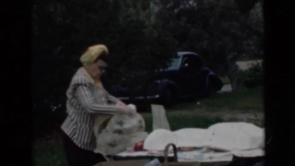 In 1949 San Diego home-movie footage, a woman in a striped blouse and yellow headscarf sets up picnic coverings near trees and a dark car.