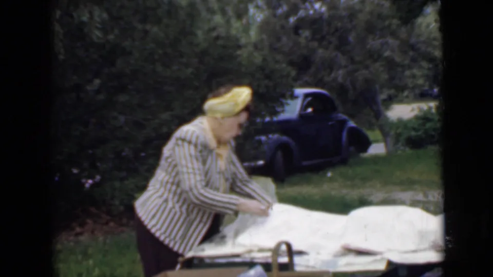 A woman continues setting up an Orange County Park picnic table in 1949 home-movie footage, with greenery and a dark car in the background.