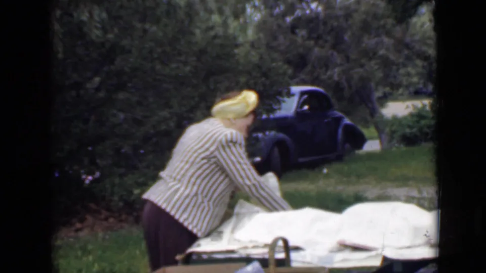The woman reaches across the picnic setup in 1949 San Diego park footage, with white coverings, greenery, and a dark rounded car visible.