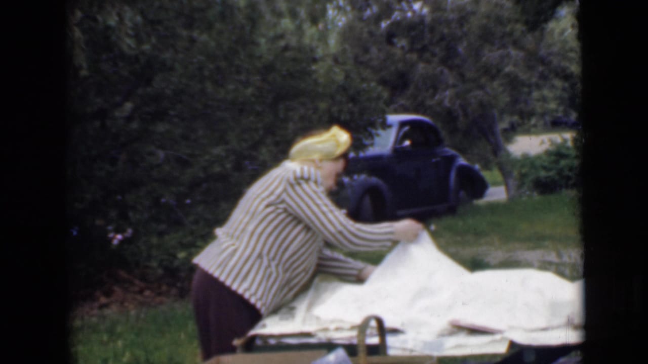 In 1949 San Diego home-movie footage, a woman in a striped blouse and yellow headscarf arranges a white picnic blanket near a dark vintage car.