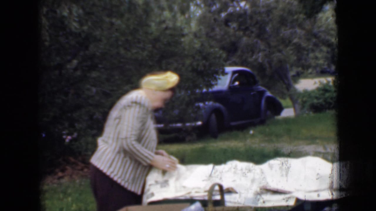 Woman bends over a picnic table with a white blanket as a dark vintage car is parked behind her.