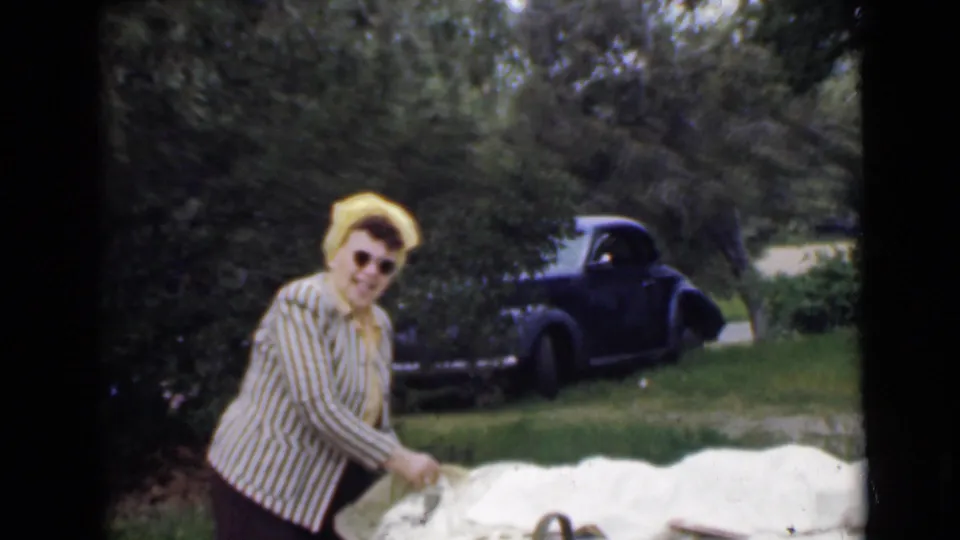 A woman in sunglasses, a striped blouse, and a yellow headscarf looks toward the camera during a 1949 San Diego park picnic scene.