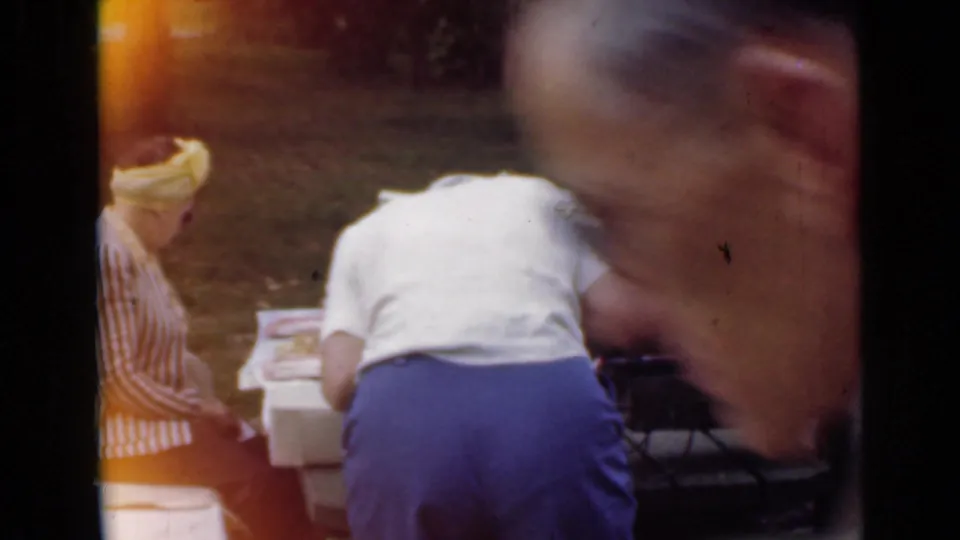 The picnic scene shifts to adults around the table, with a close foreground view of a man's face partially obscuring the 1949 San Diego park setting.