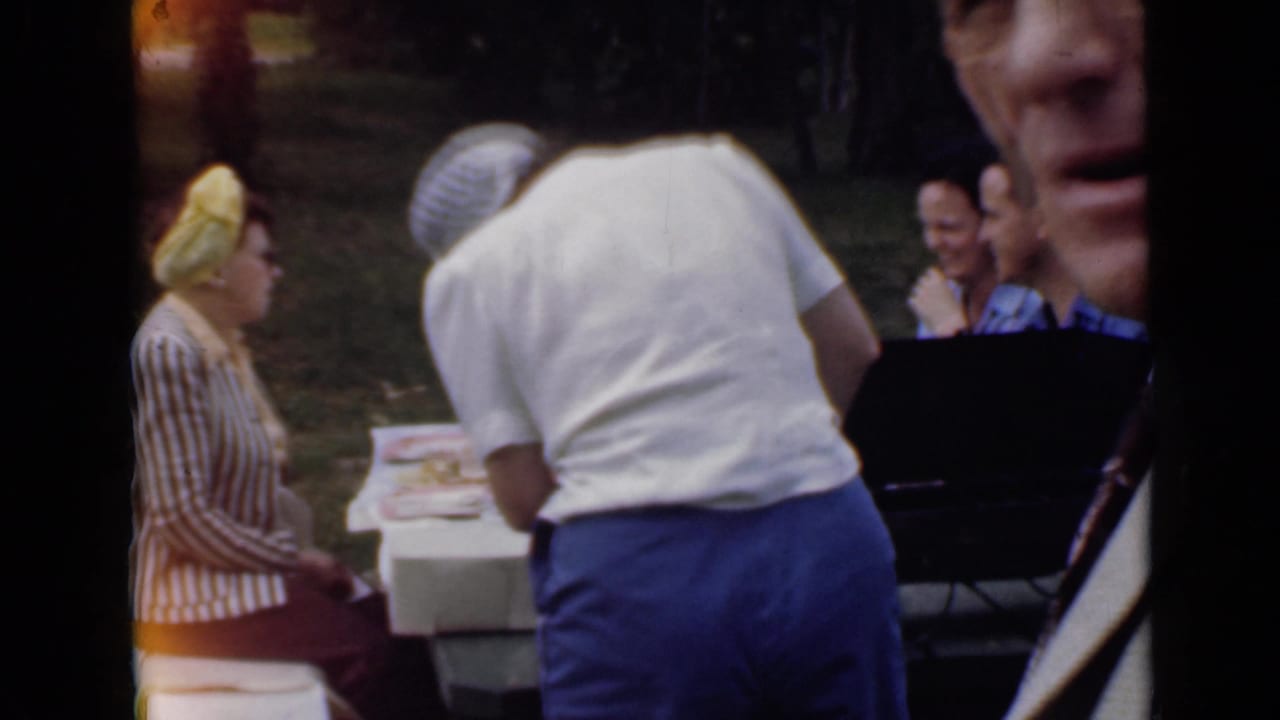 1949 San Diego home-movie frame showing adults near a white picnic table in a grassy park, including a woman in a yellow headscarf.