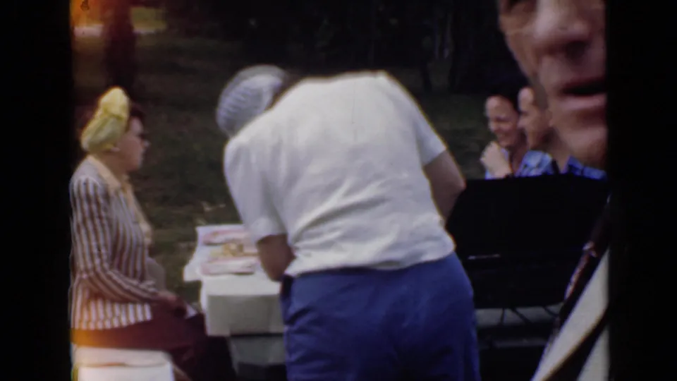 1949 San Diego home-movie frame showing adults near a white picnic table in a grassy park, including a woman in a yellow headscarf.