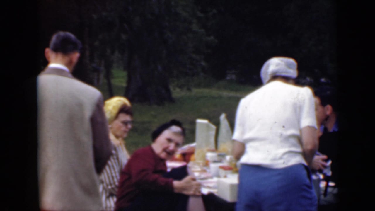 1949 San Diego outdoor picnic scene with adults in period clothing seated and standing around a white table set with food.