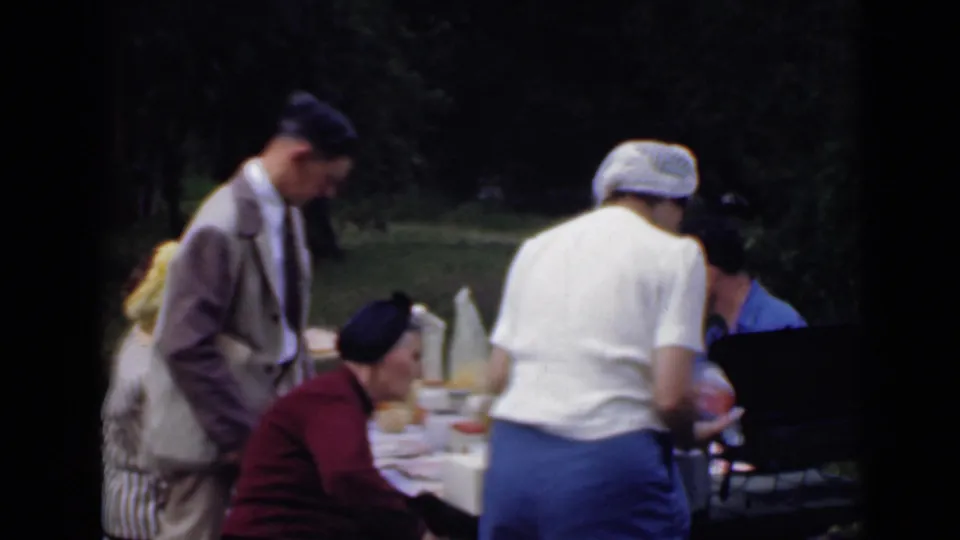1949 San Diego park picnic frame with adults seated and standing around a table, filmed with soft 8mm color and handheld framing.