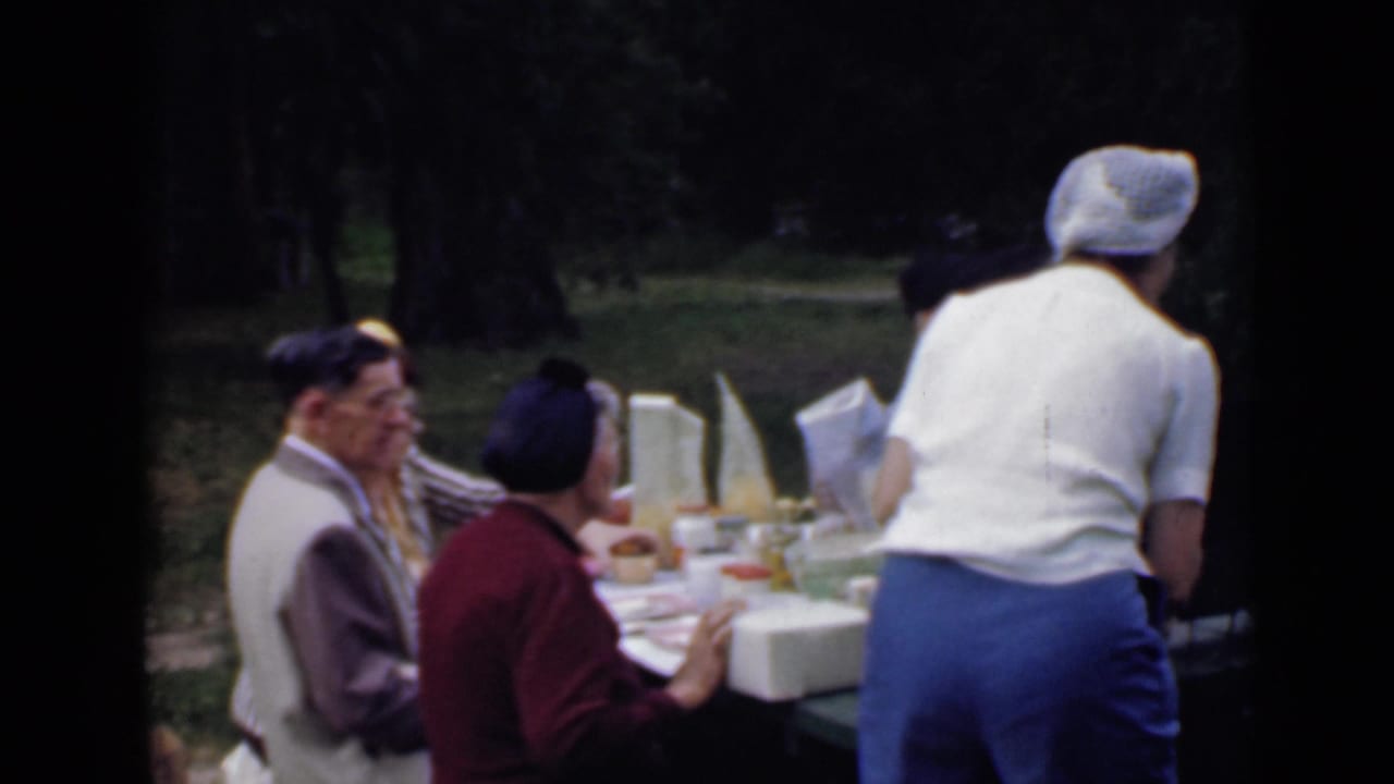 1949 San Diego home-movie frame of adults at a park picnic table with food, drinks, and a woman standing to serve.