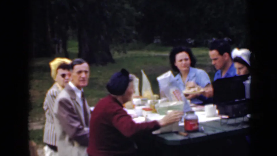 1949 San Diego home-movie frame showing adults in period clothing at an outdoor picnic table in a wooded park setting.