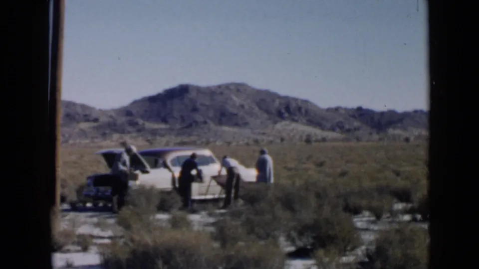 Several adults gather beside a white mid-century sedan in a dry, brushy landscape near San Diego, with distant rocky hills behind them.