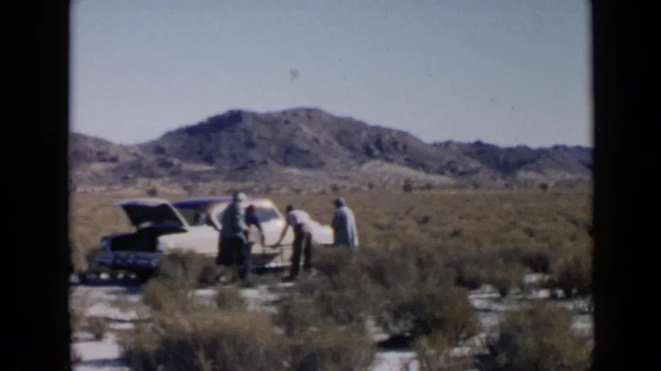 A small group stands around a white car with an open trunk or hood in a dry Southern California landscape, suggesting a roadside stop.
