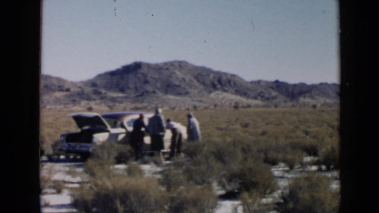 1949 San Diego-area home-movie frame showing four adults near an older car with an open trunk in arid desert scrub.